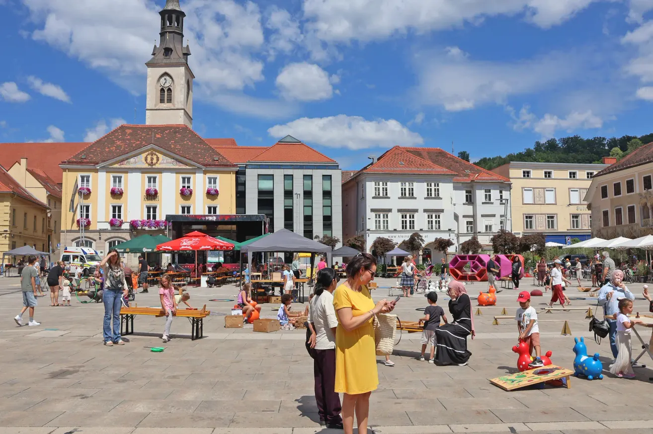 A town square with several buildings, a clock tower, and people engaging in various activities. There are tables, chairs, and umbrellas set up, and children are playing on the ground.