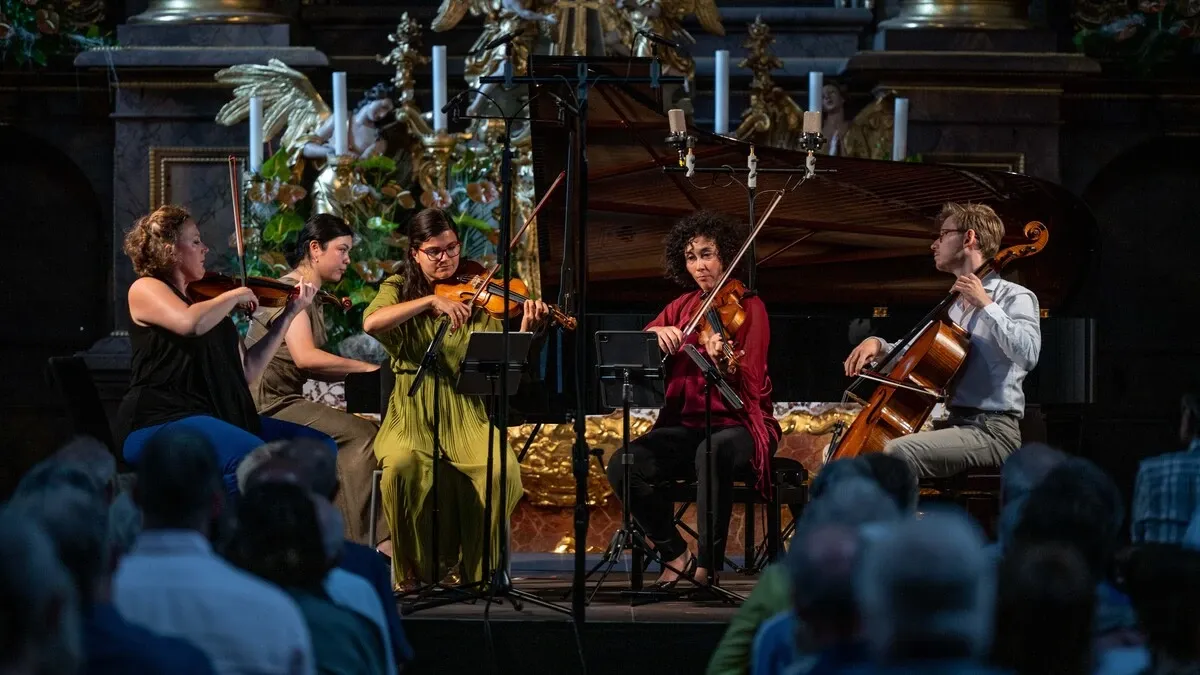 A string quartet performs on stage in front of an audience, with two violinists, a cellist, and a pianist. The musicians are dressed in casual attire and are playing in front of a golden altar.