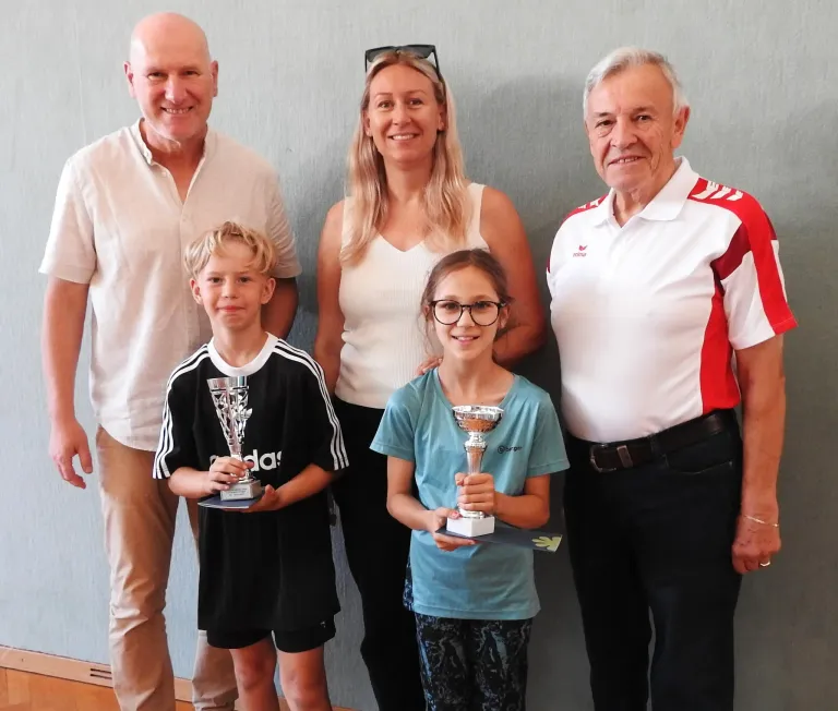 Eine vierköpfige Familie steht zusammen und hält Pokale in der Hand, mit einem lächelnden Jungen, einem Mädchen mit Brille und zwei älteren Erwachsenen, einer in einem weißen Hemd und der andere in einem rot-weißen Hemd.