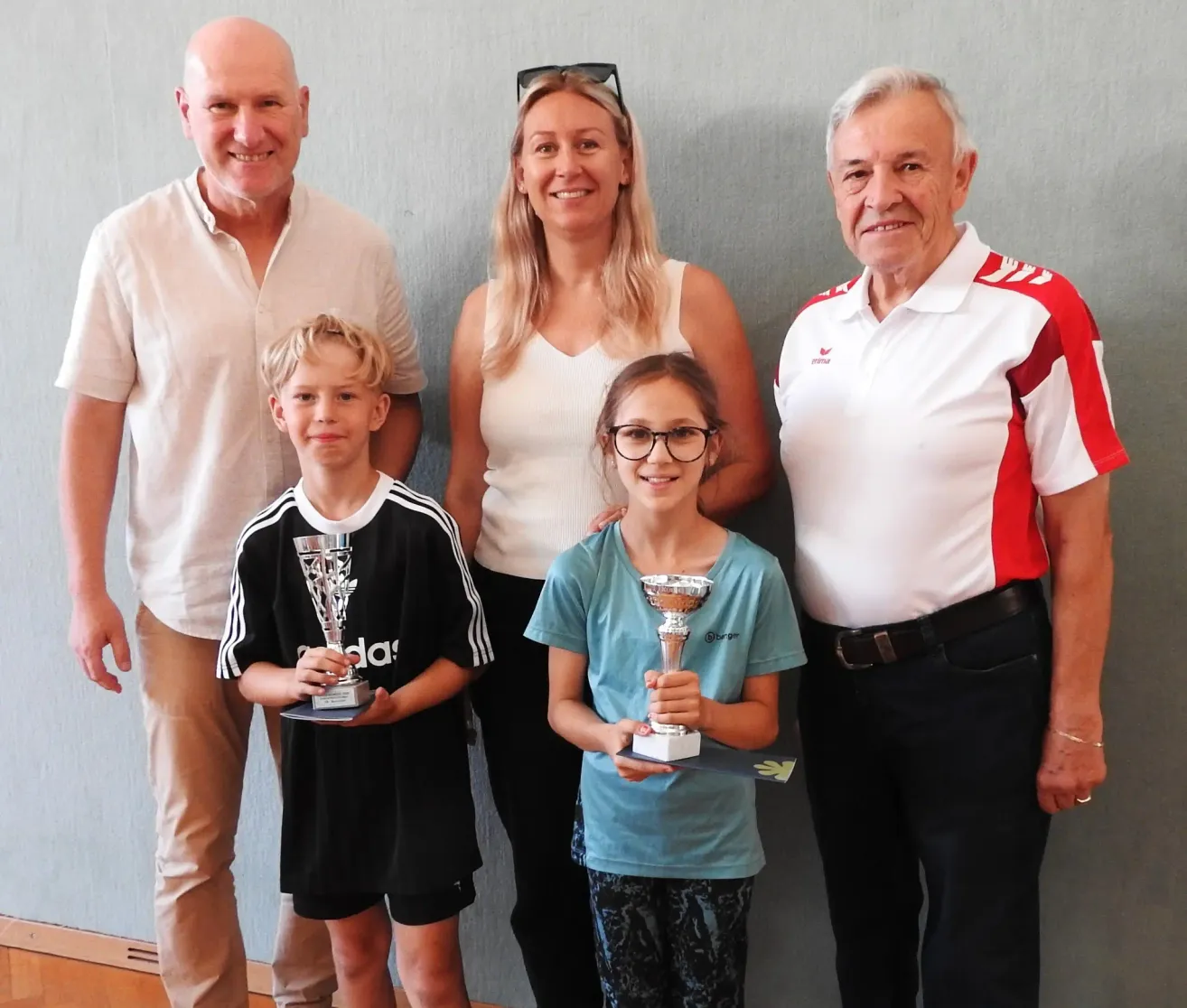 A family of four stands together holding trophies, with a smiling boy, a girl with glasses, and two older adults, one in a white shirt and the other in a red and white shirt.