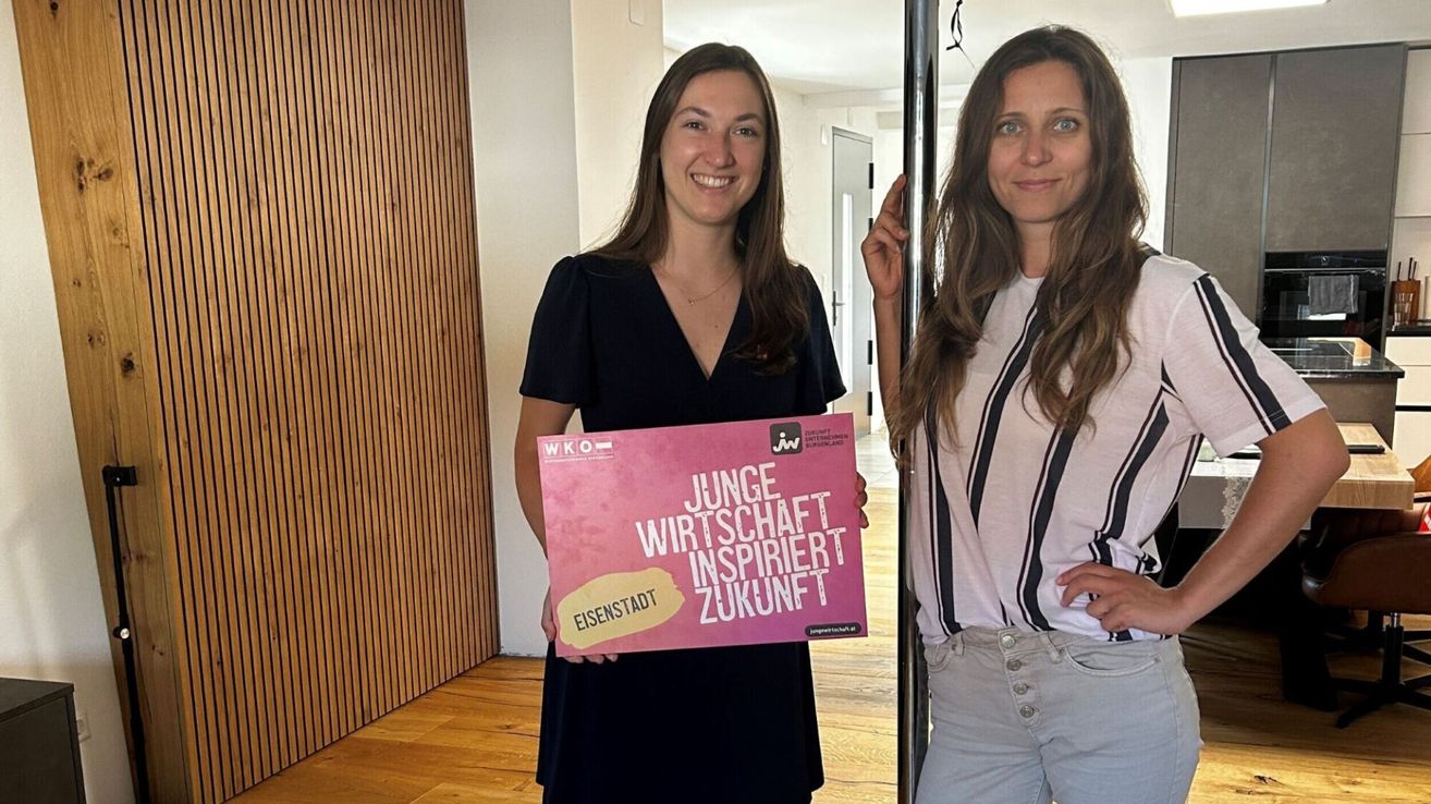 Zwei Frauen lächeln und halten ein rosa Schild mit der Aufschrift 'Junge Wirtschaft inspiriert Zukunft' in einem Raum.