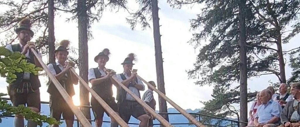 Four men in traditional attire play long wooden horns in a forest setting, with trees and a mountain backdrop.