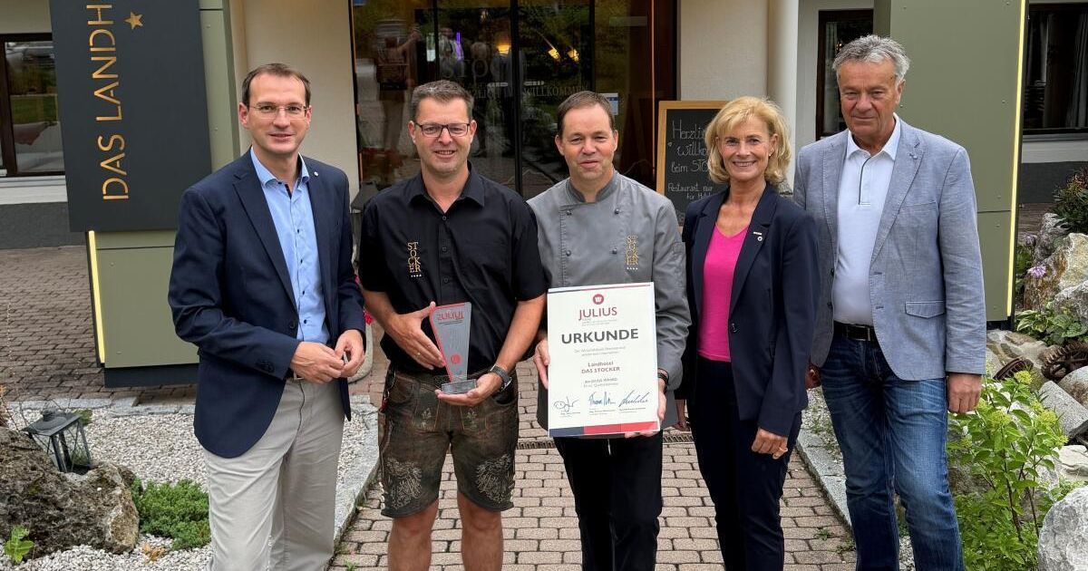 Fünf Menschen stehen vor einem Gebäude und lächeln für ein Foto. Ein Mann in der Mitte hält ein Zertifikat und eine Trophäe. Dahinter zeigt eine Tafel Text.