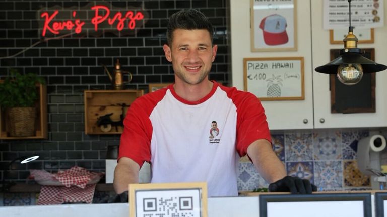 A man in a red and white shirt stands behind a counter, smiling. Behind him, a sign reads 'Pizza Hawaii' and a price tag. A QR code is visible on the counter.