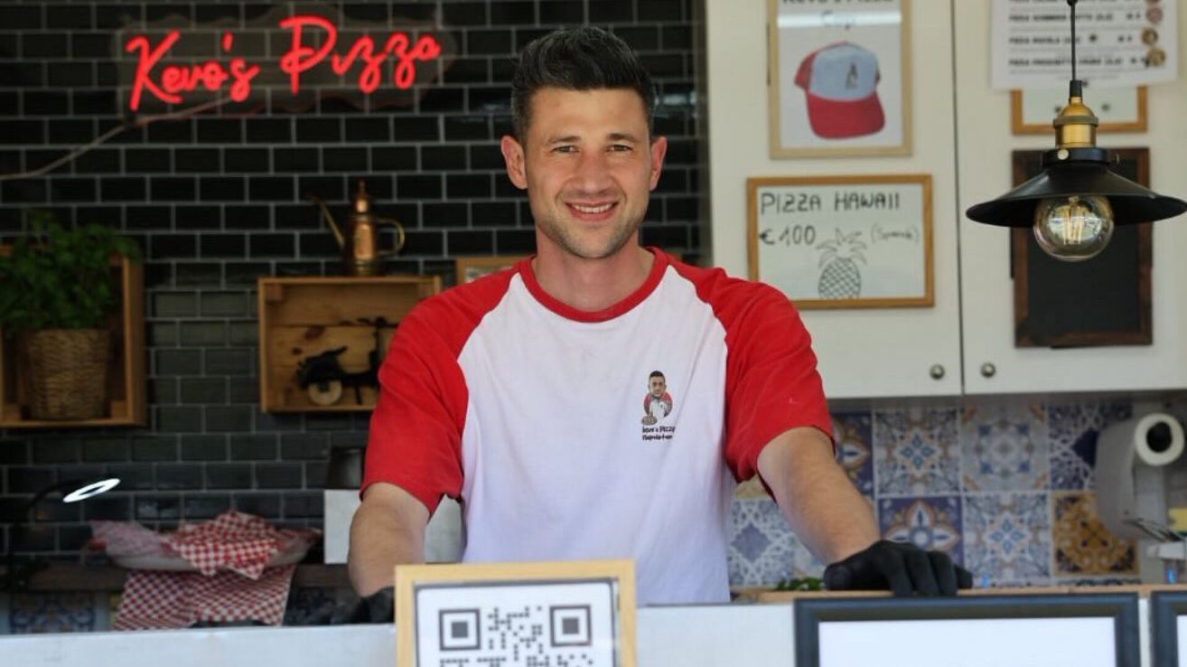 A man in a red and white shirt stands behind a counter, smiling. Behind him, a sign reads 'Pizza Hawaii' and a price tag. A QR code is visible on the counter.