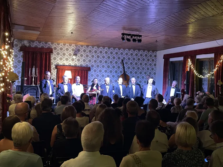 A group of musicians in suits and bow ties perform on a stage with a floral wallpaper backdrop, in front of an audience.