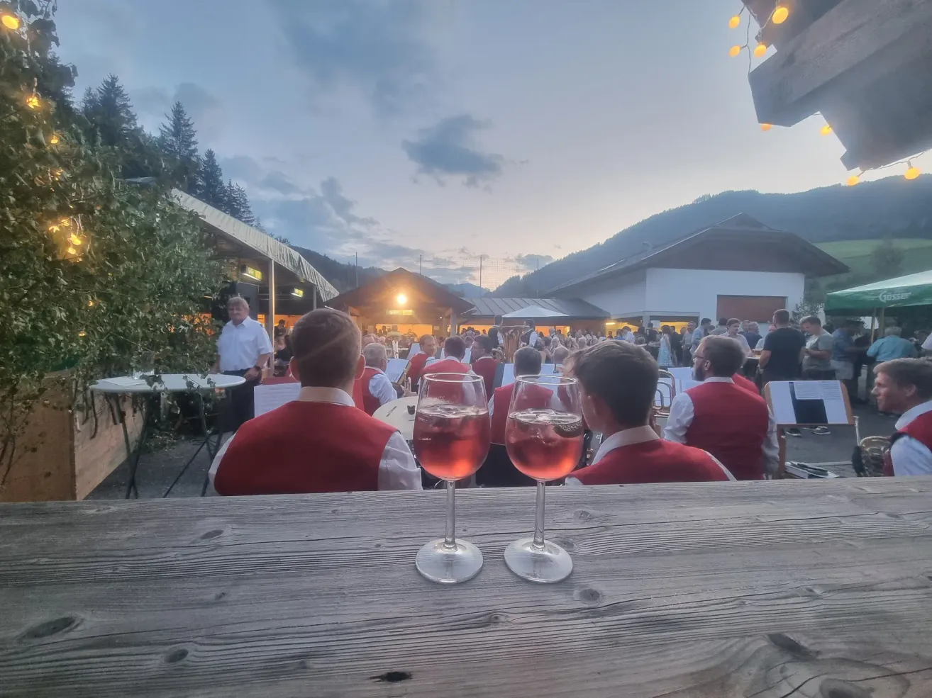 Two glasses of wine on a wooden table in front of a seated audience, with musicians in red vests performing in the background under a cloudy sky.