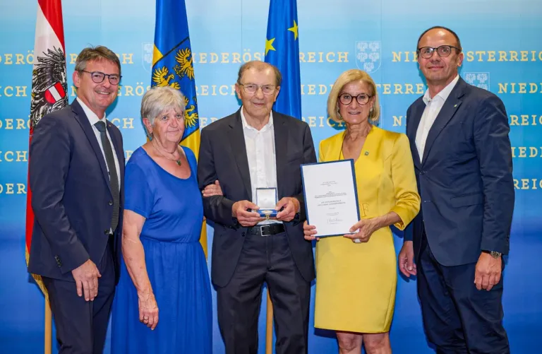 Eine Gruppe von Menschen steht zusammen und hält ein Zertifikat, mit zwei Männern und zwei Frauen, eine Frau trägt ein blaues Kleid und ein Mann in einem Anzug hält eine Medaille.