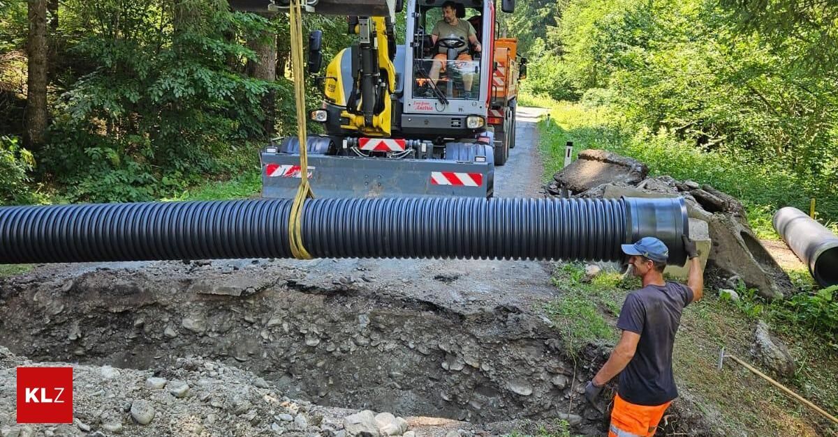 A man in an orange vest operates a large machinery vehicle, which is lifting a long black pipe over a road. Another man in a hat and gloves stands nearby.