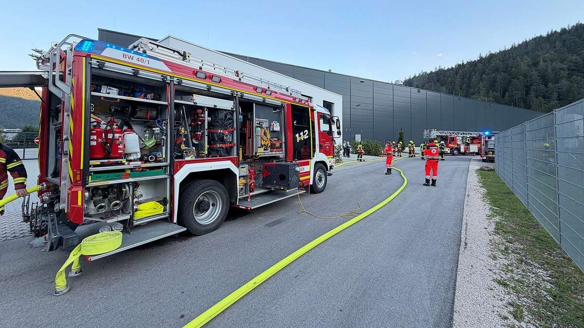 A fire truck is parked on the side of the road with its doors open, showing various firefighting equipment inside. Firefighters are standing around, some in red uniforms, with a hose laid out on the road.