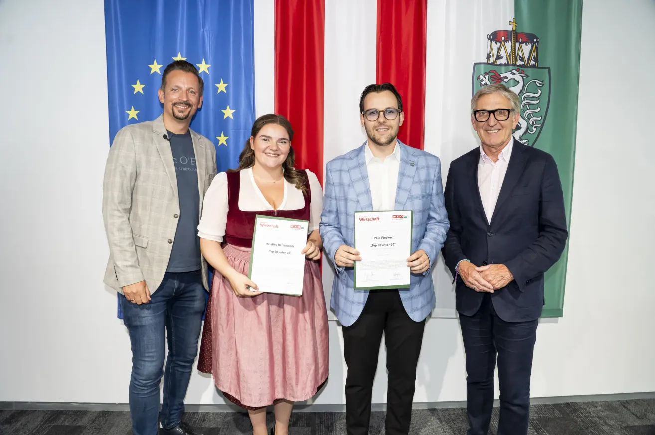 Vier Personen stehen vor Fahnen und Bannern und halten Urkunden in den Händen. Sie lächeln und scheinen sich bei einer Preisverleihung zu befinden. Der Mann rechts trägt einen Anzug und eine Brille. Der Mann in der Mitte hält eine Urkunde mit seinem Namen darauf. Die Frau links trägt ein traditionelles deutsches Kleid.