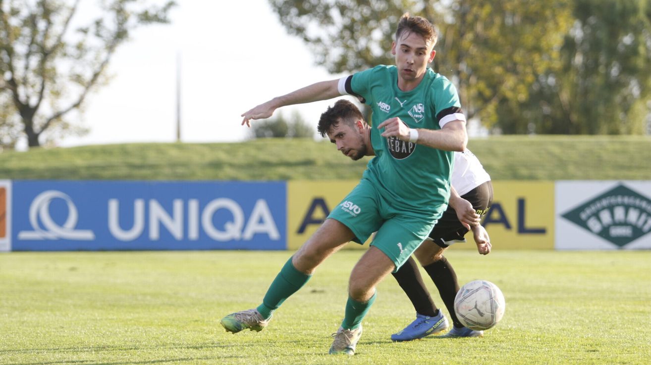 Ein Fußballspieler in einem grünen Trikot, mitten im Schuss, jagt dem Ball hinterher, während ein anderer Spieler in schwarzen Shorts versucht, ihn zu blockieren. Dahinter steht ein blaues Banner mit der Aufschrift 'UNIQA'.