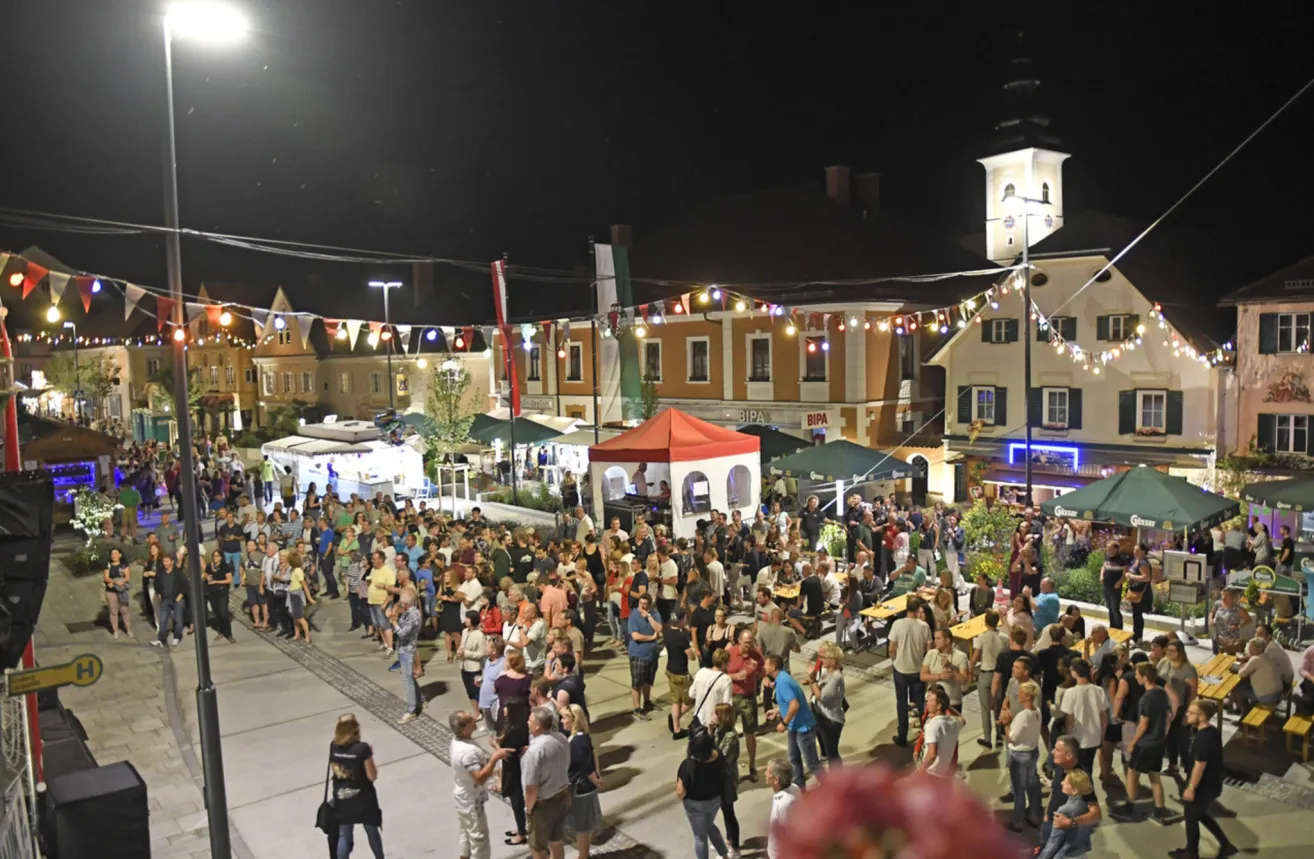 Ein lebhaftes Nachtmarkt in einem Stadtplatz, mit vielen Menschen, Zelten und Tischen. Die Gebäude haben Lichter und Flaggen, und der Himmel ist dunkel.