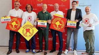 Fünf Männer stehen gemeinsam und halten Schilder in der Hand. Sie tragen weiße, grüne und schwarze T-Shirts. Jeder hält ein gelbes und rotes Schild. Der Hintergrund zeigt eine rote Kulisse mit Logos.