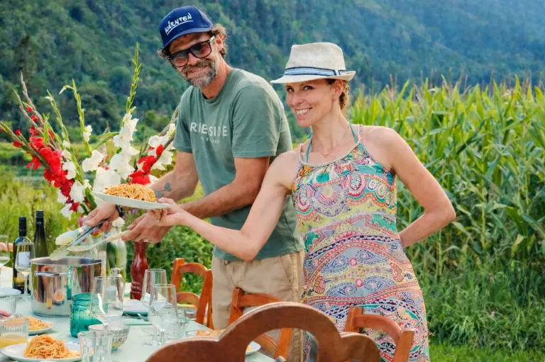 A man in a green shirt and a woman in a floral dress are standing outside, holding a plate of pasta. They are surrounded by a table with glasses and a vase with flowers. In the background, there is a field with tall grass.