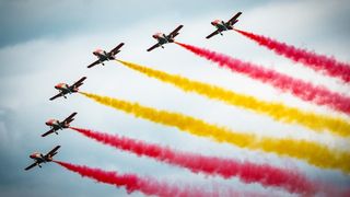 Fünf Flugzeuge mit roten, gelben und rosa Rauchspuren fliegen in Formation über einem bewölkten Himmel.