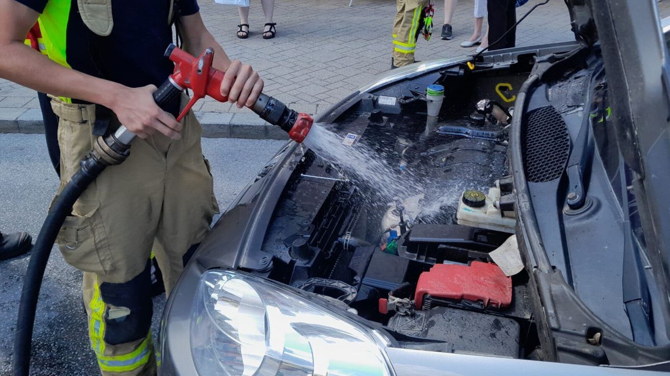 Eine Person mit einem roten Schlauch spritzt Wasser auf den Motor eines Autos, während Feuerwehrleute zusehen. Die Motorhaube ist geöffnet und der Motor ist freigelegt. Es befinden sich Flaschen und andere Gegenstände im Inneren.