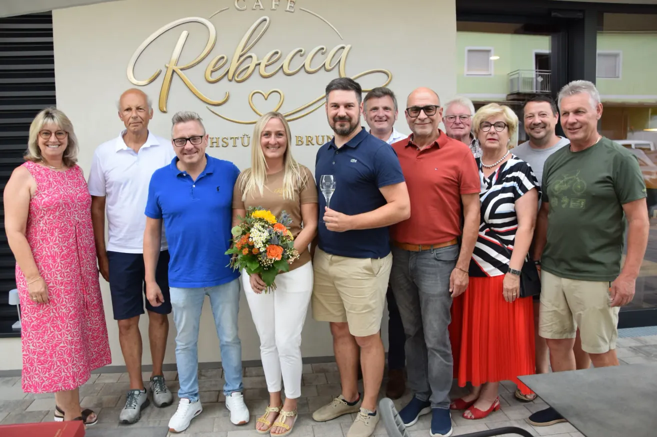 Eine Gruppe von Menschen steht vor einem Café-Schild und posiert für ein Foto. Eine Frau hält Blumen und ein Glas Champagner. Männer und Frauen sind lässig gekleidet.