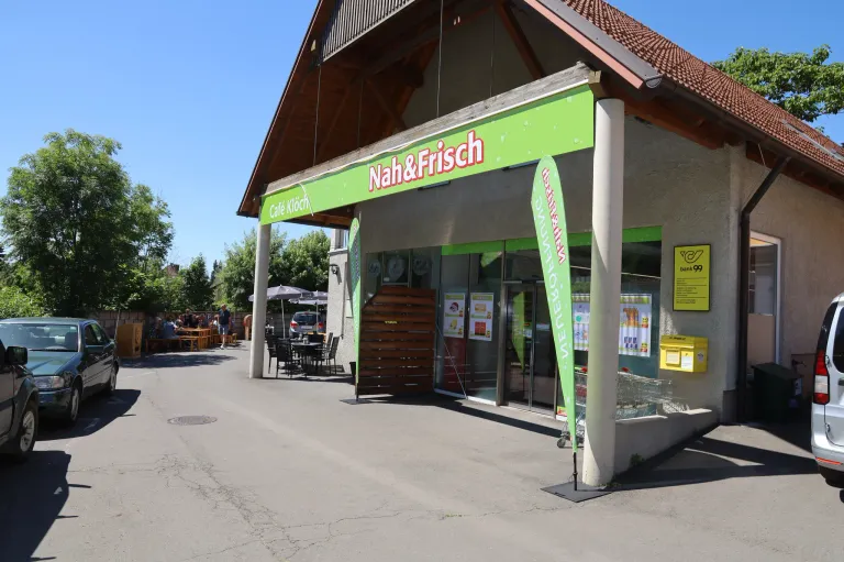 Die Außenansicht eines Cafés und Restaurants mit dem Namen Nah&Frisch. Es gibt Tische und Stühle draußen. Eine Postbox ist an der Wand.