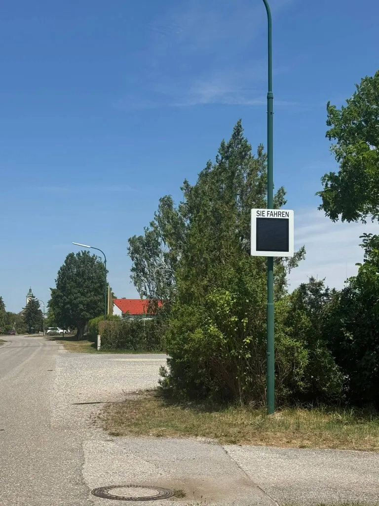 Ein Straßenschild auf einem Pfosten weist Autofahrer an, zu fahren. Eine Straßenlaterne steht auf der linken Straßenseite, während Bäume und Büsche beide Seiten säumen.