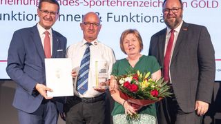 Ein Mann und eine Frau stehen neben zwei Männern, alle halten eine Auszeichnung, Blumen und ein Zertifikat. Sie sind auf einer Bühne, lächeln und posieren für ein Foto. Der Banner hinter ihnen lautet 'Bundes-Sportehrenzeichen'.