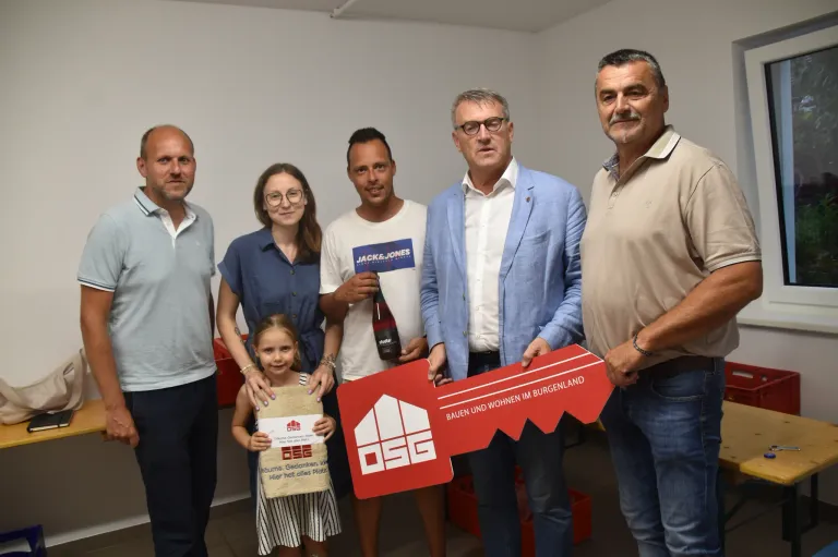 Six people stand together with a key prop in a room. The girl holds a small box. The man on the right wears a watch. The man on the left wears glasses. The man in the middle holds a bottle.