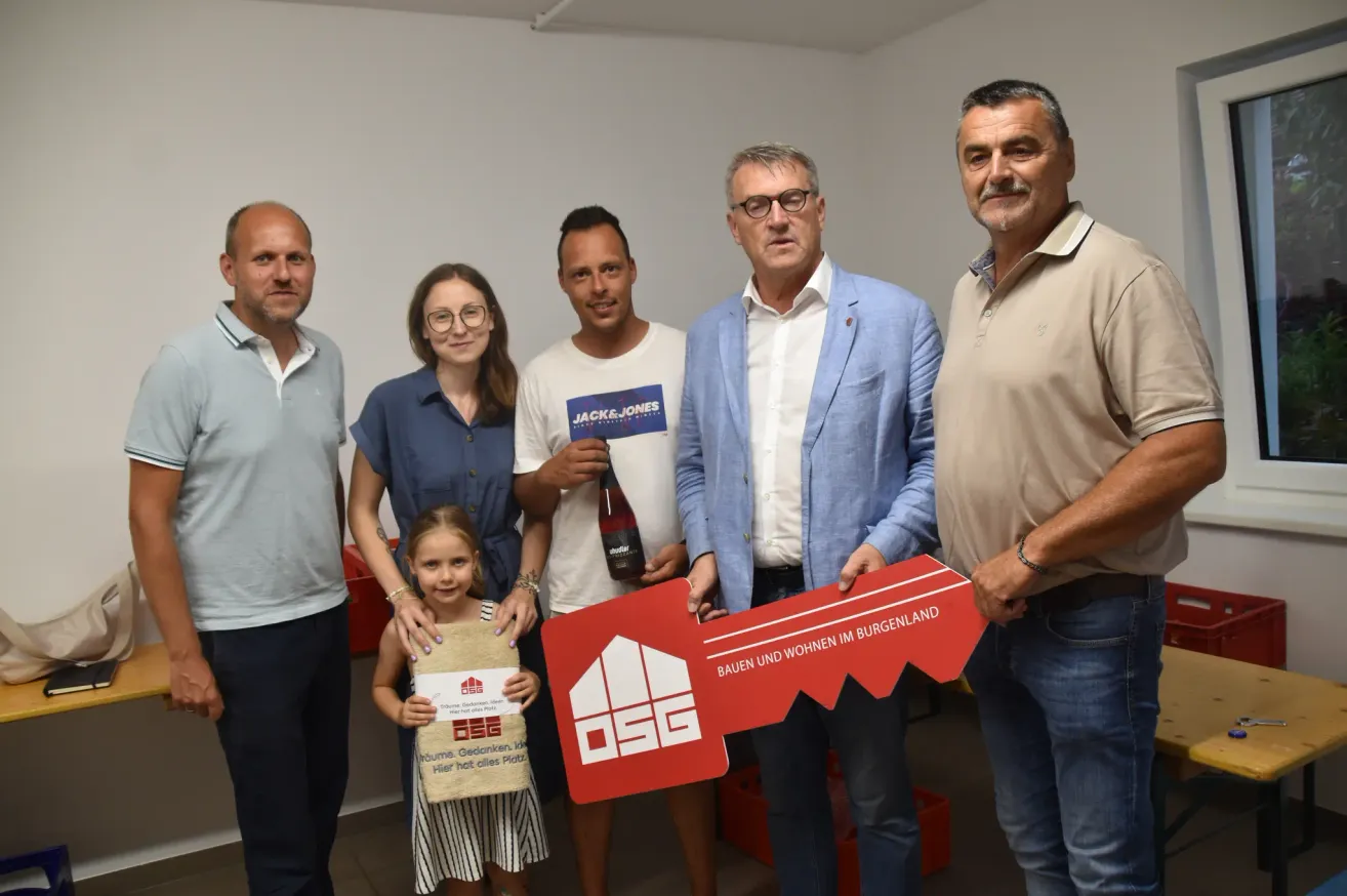 Six people stand together with a key prop in a room. The girl holds a small box. The man on the right wears a watch. The man on the left wears glasses. The man in the middle holds a bottle.