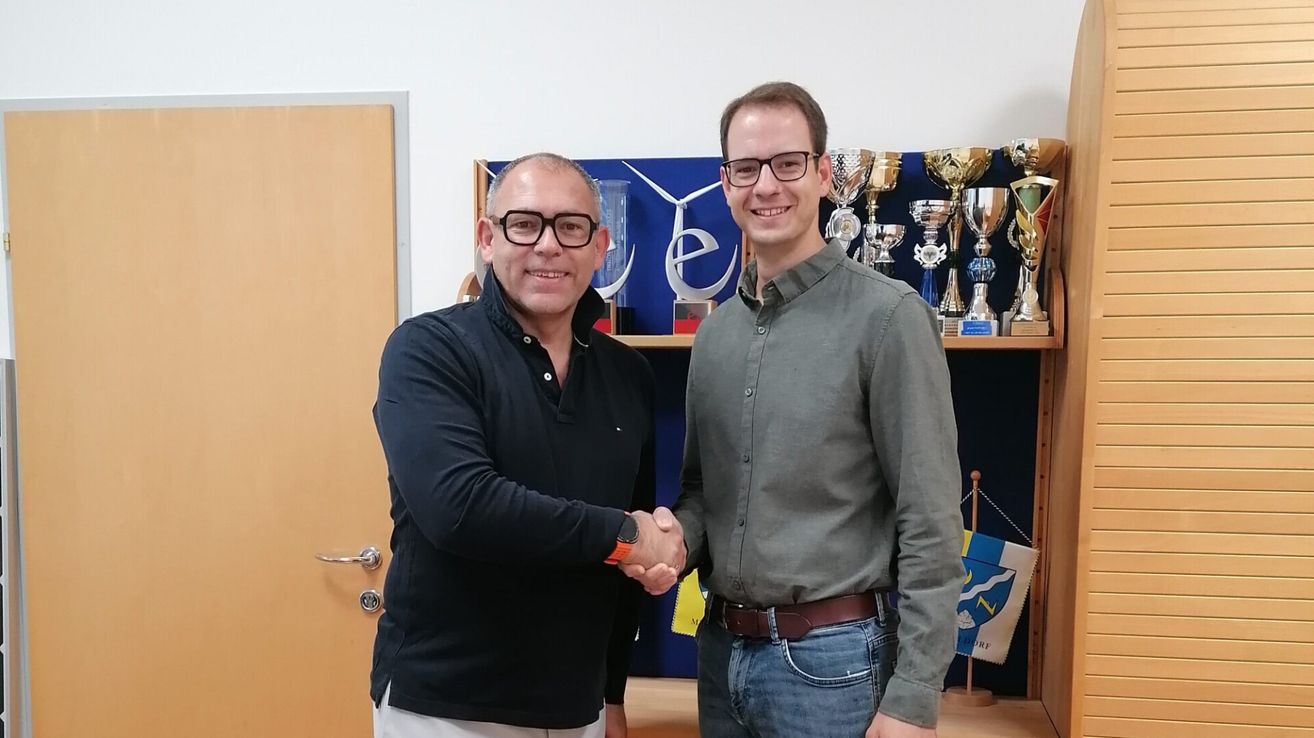 Zwei Männer stehen lächelnd und schütteln sich die Hände vor einem Regal mit Trophäen und einer Flagge. Der Mann auf der linken Seite trägt ein schwarzes Shirt, während der Mann auf der rechten Seite ein graues Shirt und eine Brille trägt.