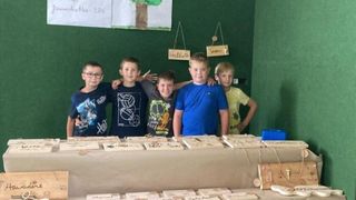Fünf Jungen stehen hinter einem Tisch mit geschnitzten Holztafeln. Hinter ihnen ist eine grüne Wand mit drei Holzschildern. Eines heißt 'Sennas', ein anderes 'Hallo' und das dritte ist leer.