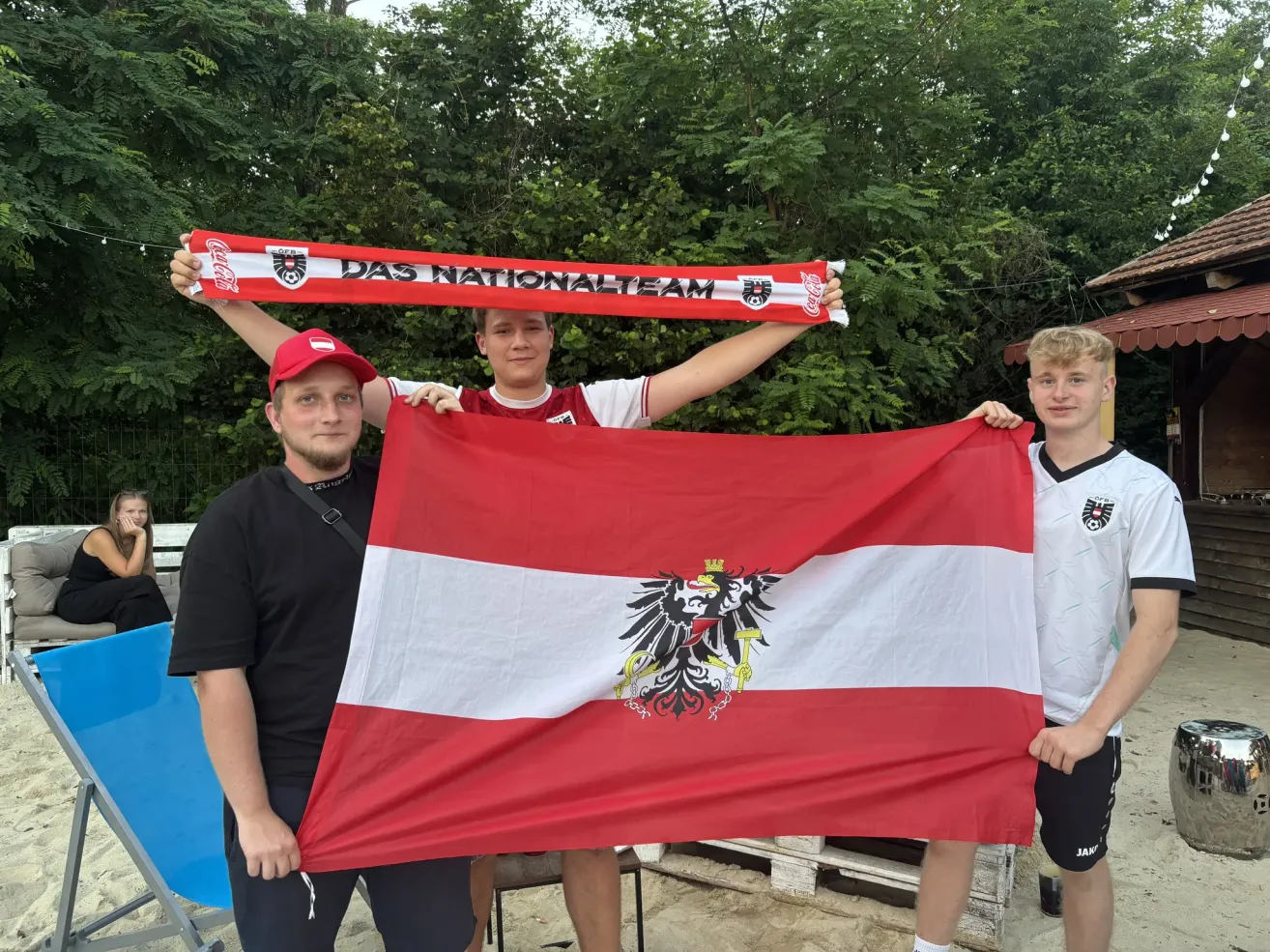 Drei Männer halten eine rot-weiße Flagge mit einem Adler und einem Banner mit der Aufschrift 'DAS NATIONALTEAM'. Sie stehen vor Bäumen.