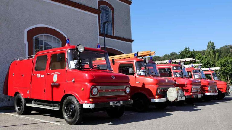 Several vintage red fire trucks are parked outside a building with large windows. They have blue lights on top and are parked in a row.