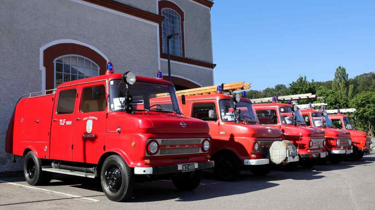Several vintage red fire trucks are parked outside a building with large windows. They have blue lights on top and are parked in a row.