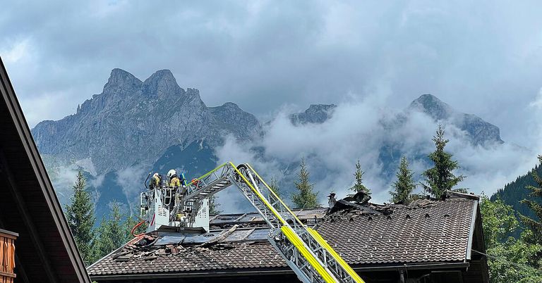 Firefighters on a ladder are working on a roof damaged by fire with mountains in the background.