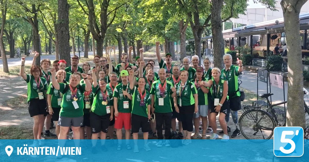 A group of people in green uniforms with medals pose for a photo under trees. They are smiling and raising their hands.