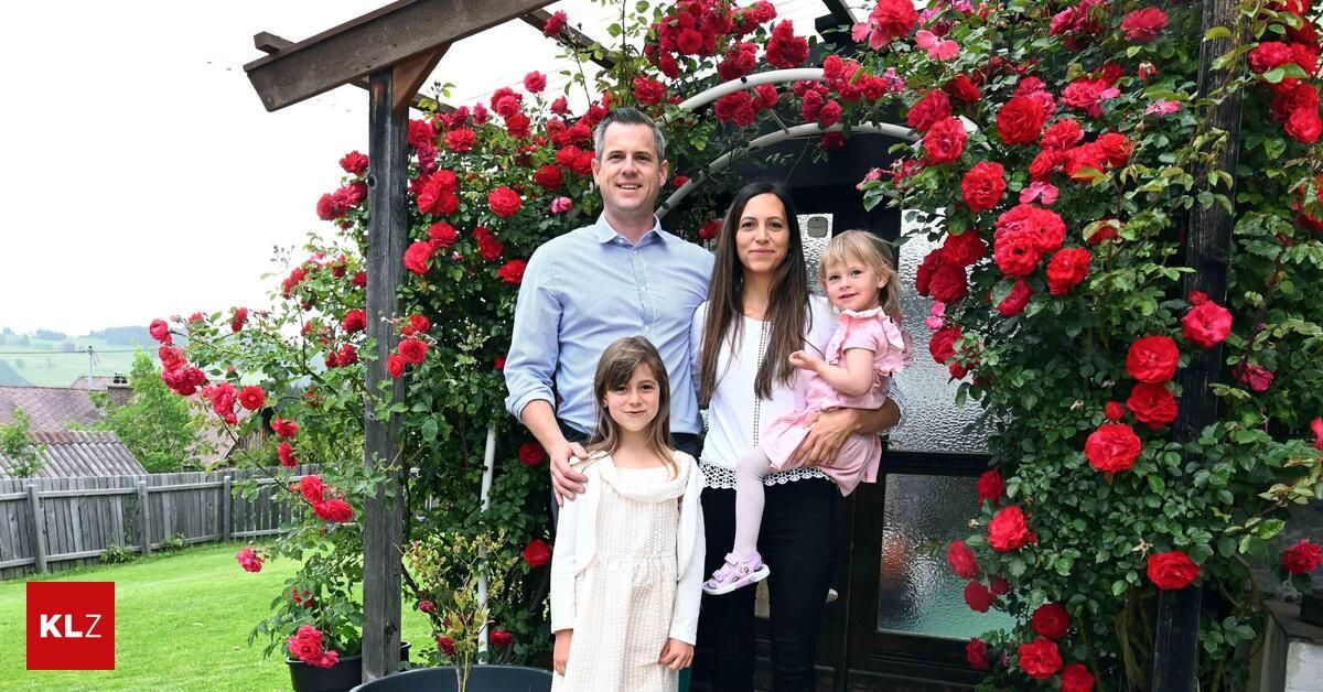 Eine Familie steht vor einem Rosengarten und lächelt für das Foto. Der Mann hält ein Kleinkind auf seiner linken Seite, während die Frau ein weiteres Kind auf der rechten Seite hält. Hinter ihnen ist der Garten mit blühenden roten Rosen gefüllt.