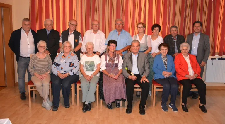 A group of older adults are sitting together in rows, smiling for a photo in a room with a red curtain. Some are wearing glasses, watches, and rings. A man in the middle holds a wine bottle.