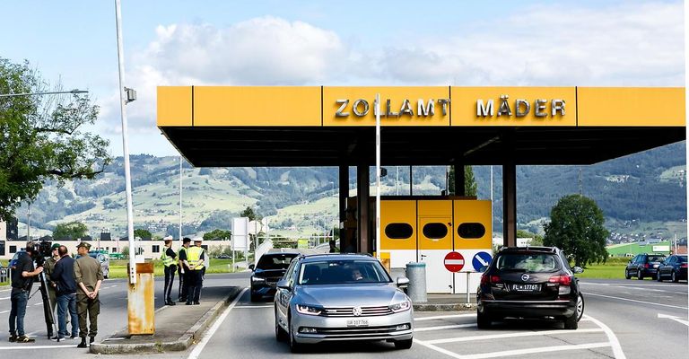 A toll booth in Zollamt Mader, with cars passing through. Two individuals in yellow vests stand by the booth.