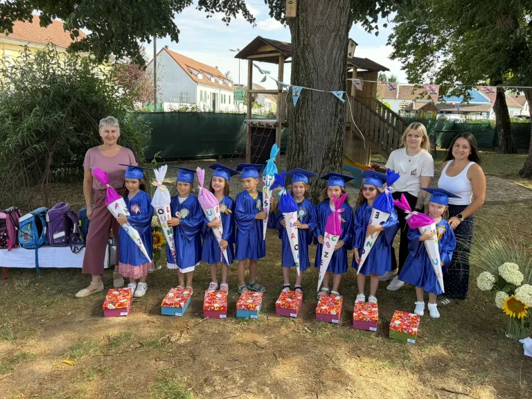 A group of young children in blue graduation gowns stand in front of a tree with decorative boxes and cones, with two women standing on the side. In the background, there are houses and a treehouse.