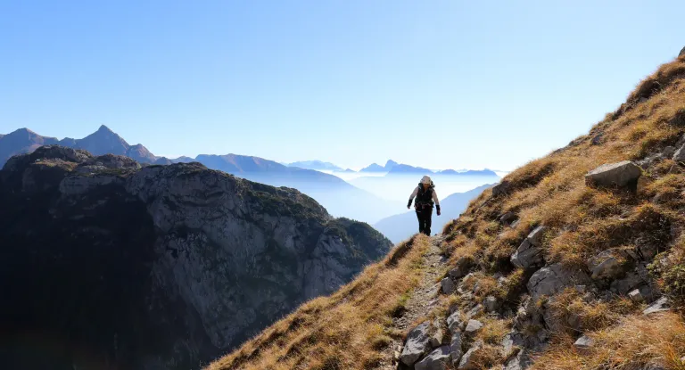 A hiker walks on a rocky mountain path with a clear blue sky and distant foggy mountains in the background.