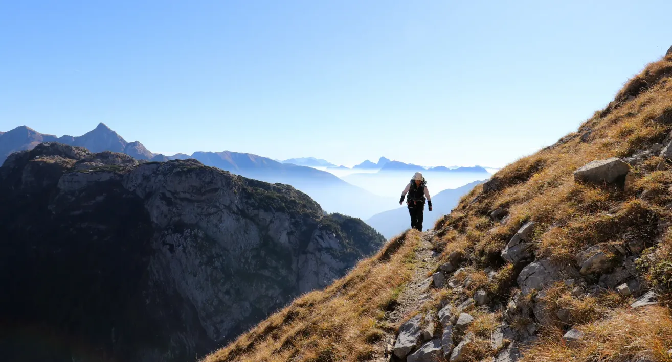 A hiker walks on a rocky mountain path with a clear blue sky and distant foggy mountains in the background.