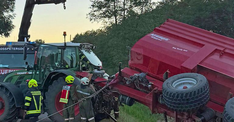 Zwei Personen in Schutzkleidung helfen bei einem umgestürzten Traktor, an dem ein roter Kipper-Anhänger beteiligt ist. Bäume und ein Sonnenuntergang im Hintergrund sind sichtbar.