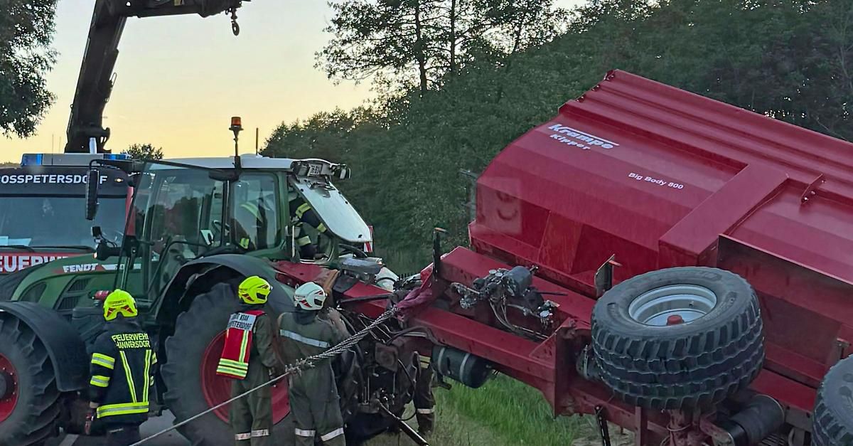 Zwei Personen in Schutzkleidung helfen bei einem umgestürzten Traktor, an dem ein roter Kipper-Anhänger beteiligt ist. Bäume und ein Sonnenuntergang im Hintergrund sind sichtbar.