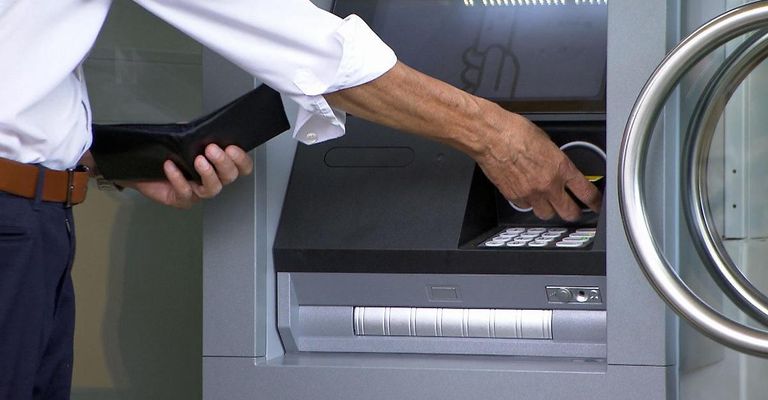 Eine Person in einem weißen Hemd benutzt einen Geldautomaten. Sie hält eine Brieftasche und steckt eine Karte in den Automaten.