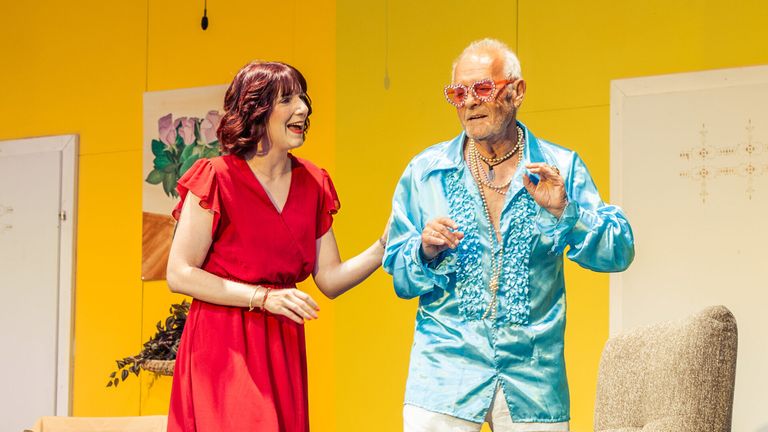 A woman in a red dress and an older man in a blue shirt with pearl necklaces are standing on a stage, possibly performing a comedy show.