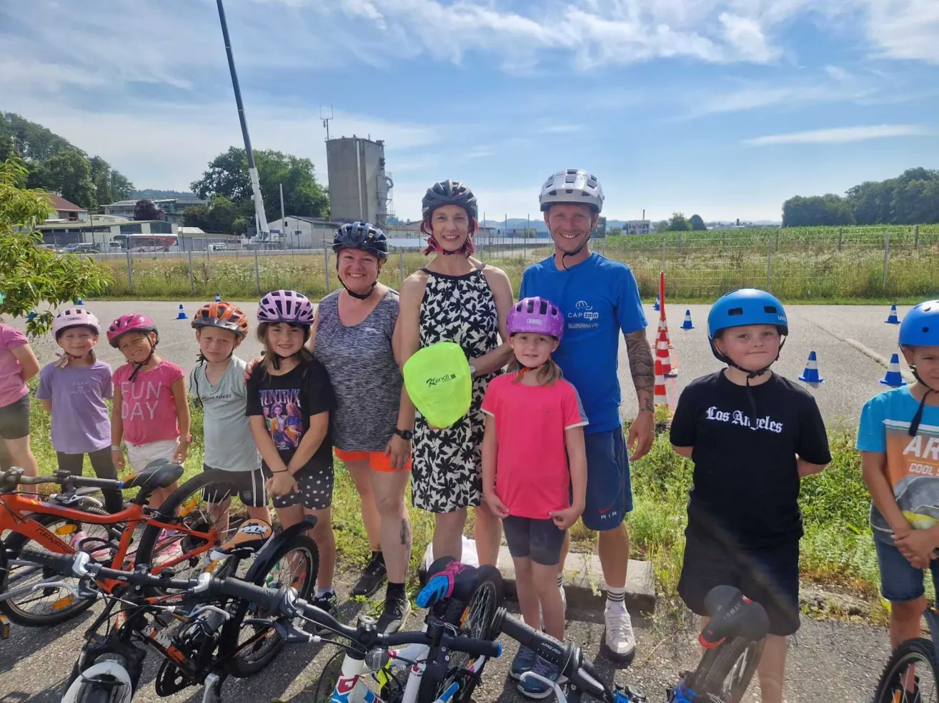 Eine Gruppe lächelnder Erwachsener und Kinder mit Helmen steht mit ihren Fahrrädern. Der Hintergrund zeigt ein Grasfeld mit einem Gebäude und einigen Baugeräten.