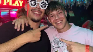 Zwei Männer in einem Club, einer mit Sonnenbrille und der andere in einem rosa Shirt, zeigen auf die Kamera. Der Hintergrund ist bunt mit Neonlichtern.