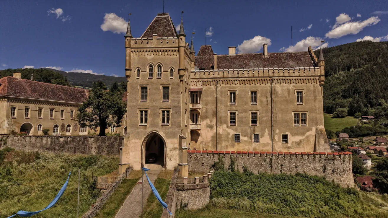 A large, aged castle with multiple turrets, set against a backdrop of mountains and blue sky with clouds. The castle features an arched entrance, multiple windows, and a stone wall in the foreground.