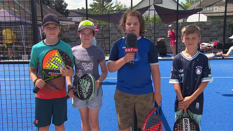 Four kids stand on a blue court, holding rackets. One boy in blue speaks into a microphone with an ORF logo. Behind them is a fence and a person in pink.