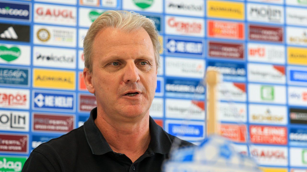 Ein Mann in einem schwarzen Poloshirt steht vor einer Wand mit verschiedenen Logos, wahrscheinlich in einer Pressekonferenzsituation.