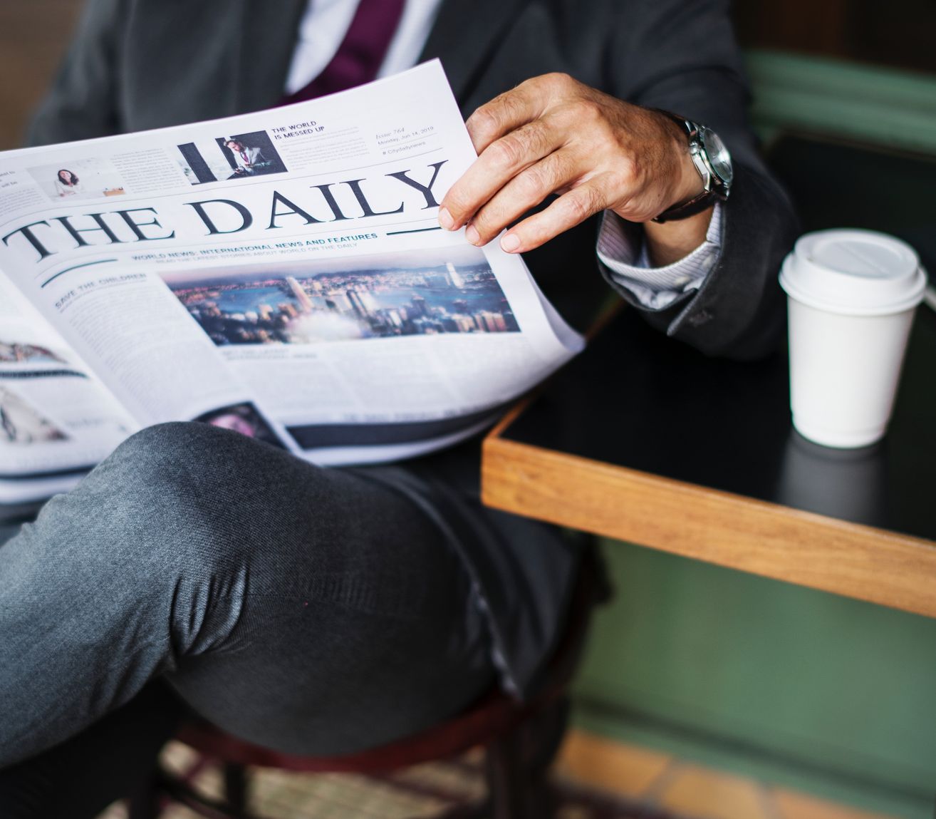 A man reads a newspaper titled The Daily while seated at a table, with a coffee cup next to him.